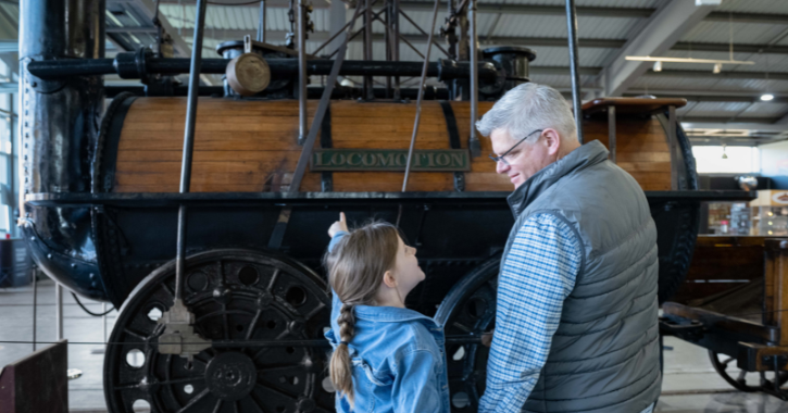 Older man and little girl stood admiring a replica of Locomotion No.1 engine at Locomotion Railway Museum.